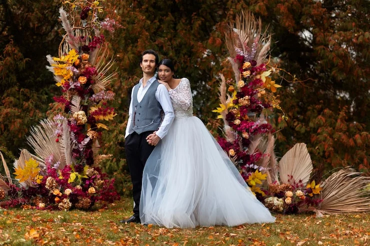 Bride in white gown and groom in vest pose outdoors among autumn floral decorations.