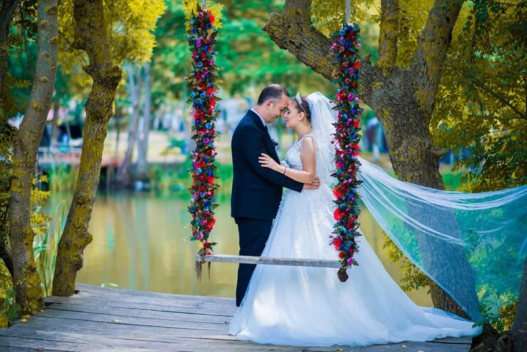 Bride and groom embracing on a decorated swing by a lake surrounded by trees in autumn.
