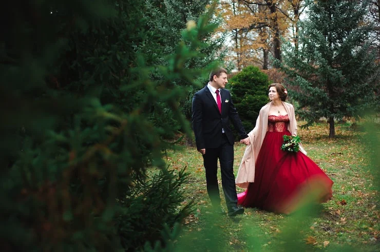 Couple in formal attire walking hand in hand through a wooded area with autumn leaves.