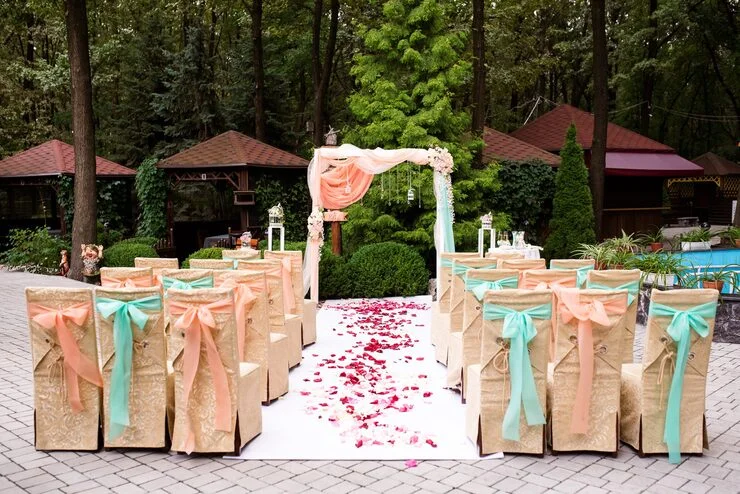 Outdoor wedding setup with beige chairs, peach and turquoise bows, white aisle, and floral arch.